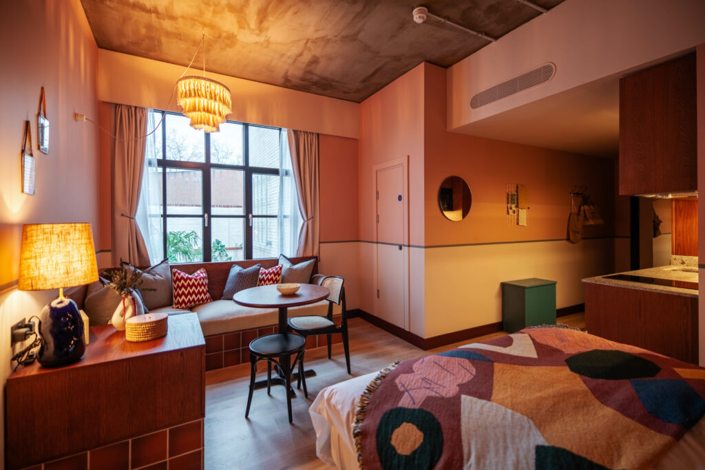 The end of a bed in the forefront with a couch and table in the rear. A hallway on the right with a kitchenette.