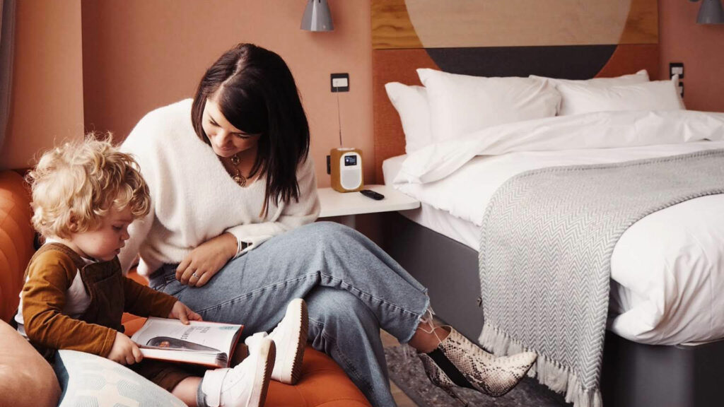 A woman and child sit on a couch reading a book in a hotel room with a large bed in the back.